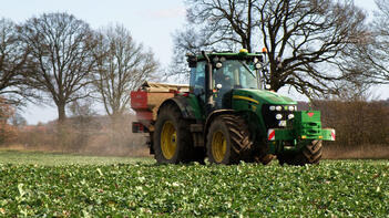 Traktor fährt über ein Feld mit grünem Bewuchs. Im Hintergrund sind Bäume und ein klarer Himmel zu sehen. (automatisch generiert durch KI)