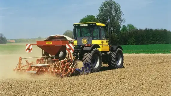 Traktor mit Anhänger pflügt ein Feld unter blauem Himmel. Im Hintergrund sind Bäume und grüne Wiesen zu sehen. (automatisch generiert durch KI)