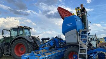 Eine Person steht auf einer Leiter neben einer großen blauen Landmaschine auf einem Feld, mit einem Traktor im Hintergrund. (automatisch generiert durch KI)