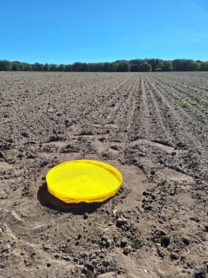 Gelbes rundes Objekt auf einem frisch gepflügten Feld vor einem Waldrand unter klarem blauem Himmel. (automatisch generiert durch KI)