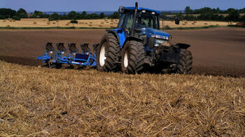 Ein blauer Traktor pflügt ein Feld, im Hintergrund sind Bäume und ein weiter Himmel sichtbar. (automatisch generiert durch KI)