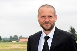 Mann in Anzug mit Krawatte steht draußen vor einem Feld und Bäumen. (automatisch generiert durch KI)