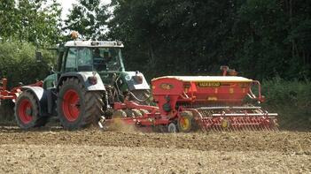 Traktor mit Sämaschine bearbeitet ein Feld, umgeben von Bäumen. (automatisch generiert durch KI)
