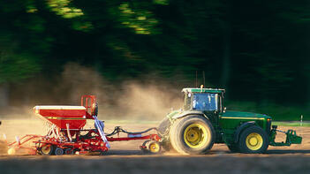 Traktor zieht landwirtschaftliches Gerät über ein Feld, im Hintergrund Bäume. (automatisch generiert durch KI)