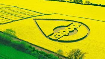 Luftaufnahme eines gelben Rapsfeldes mit einem großen Tropfenmuster und dem Wort `crop` darin. Im Vordergrund grüne Bäume und Felder. (automatisch generiert durch KI)